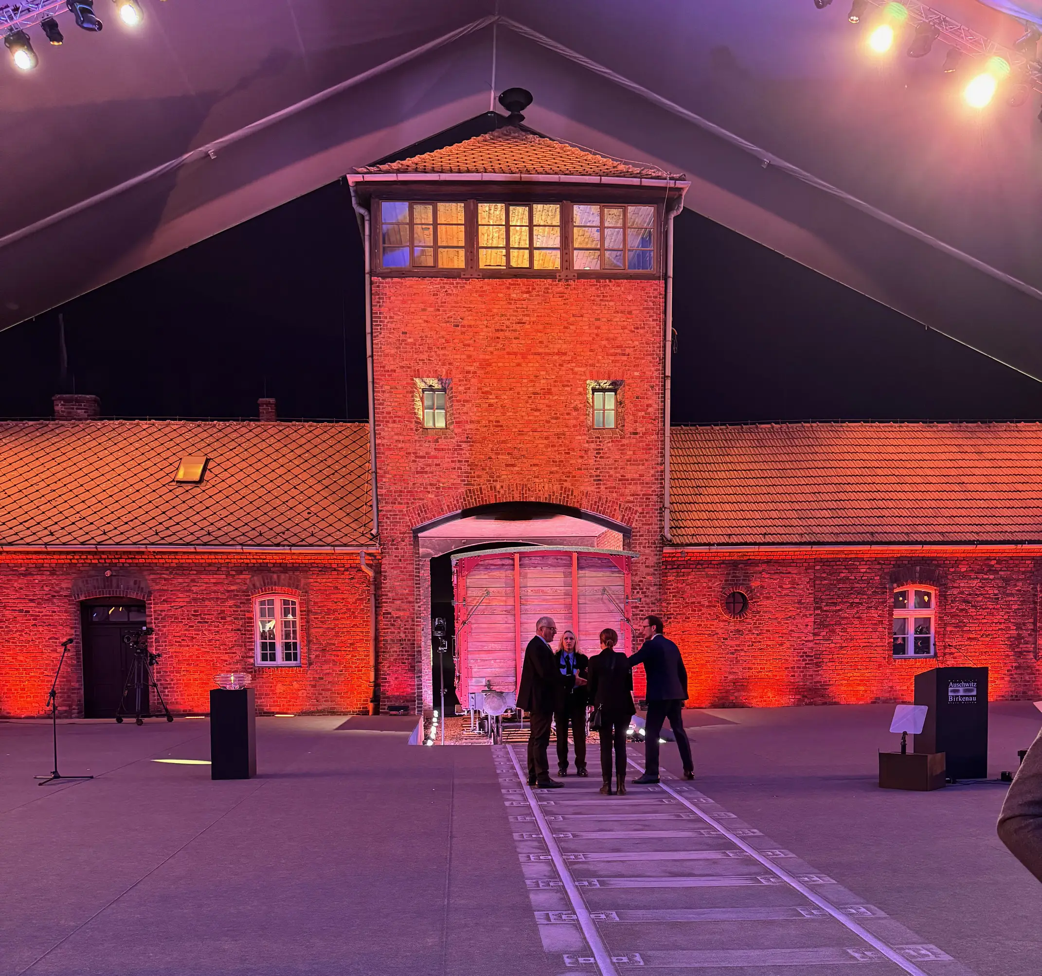 A nighttime event setup at Auschwitz's main gate, with dramatic lighting, a railway track, and a historical train car.